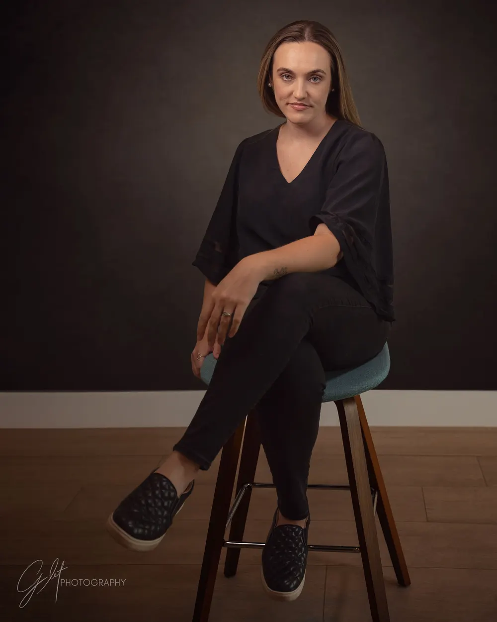 Seated on a stool, legs angled to one side, leaning with a hand resting on the knee — easy and engaged