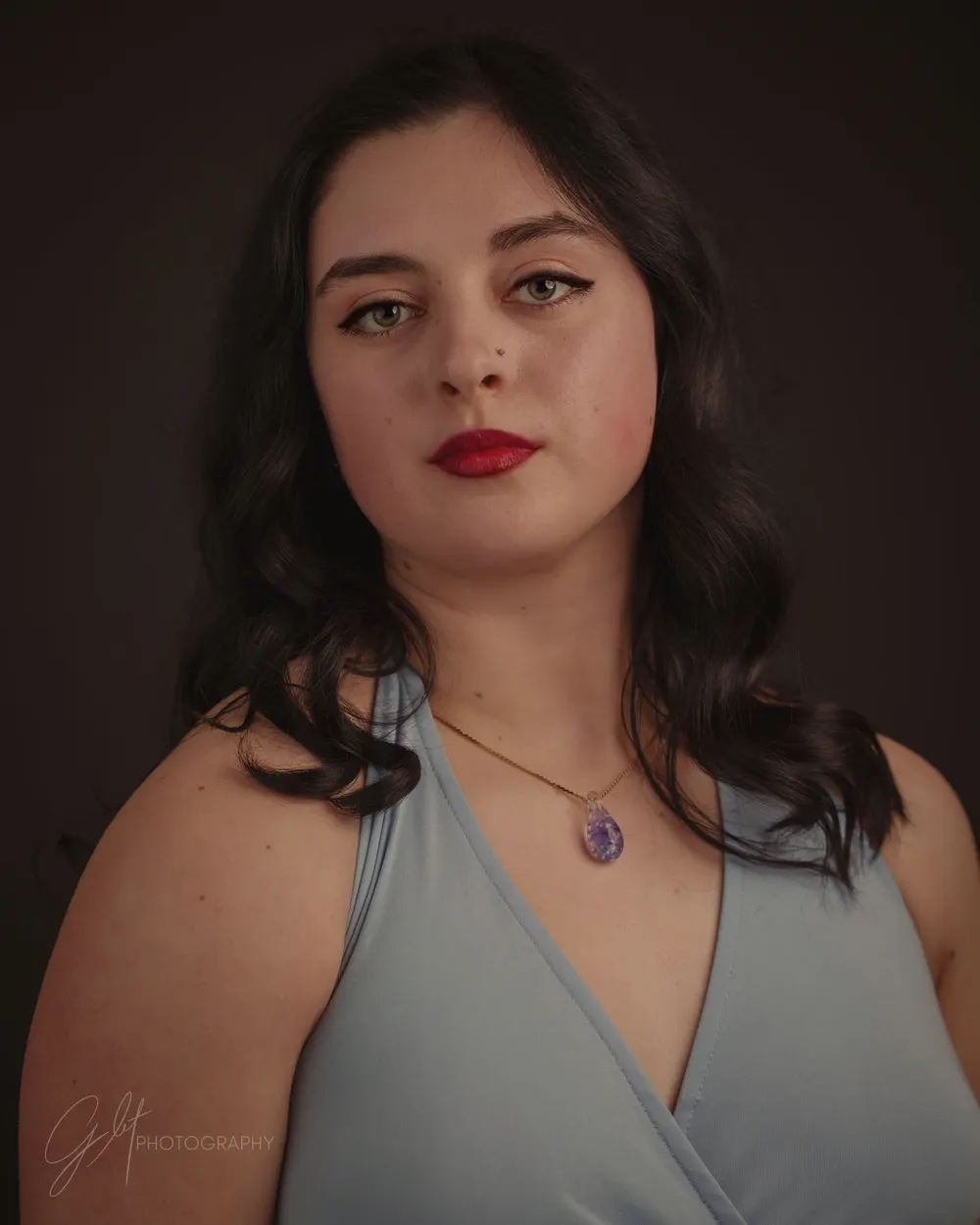 A young woman in a soft blue wrap V-neck with a simple pendant — the neckline framing her face